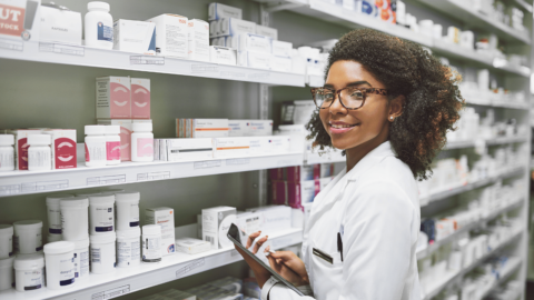 A friendly pharmacist stands in front of a pharmacy shelf filled with medications like Ozempic