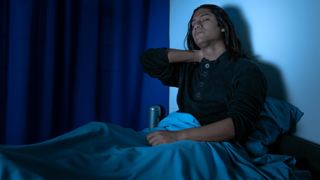 Man sitting up in bed at night, holding his neck in discomfort, representing the struggle of opioid withdrawal symptoms worsening at night and sleep difficulties during detox