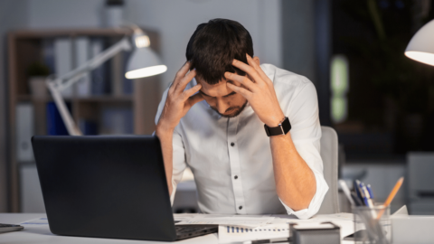 Stressed man at his desk with a laptop, holding his head in frustration, representing the challenges of balancing work and opioid addiction treatment