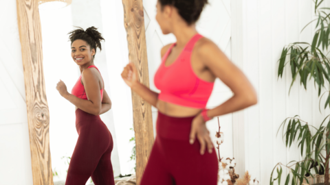 A smiling woman in workout clothes happily checks her reflection in the mirror, reflecting confidence in maintaining weight loss after using GLP-1 medications